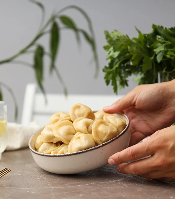 Hands holding a bowl of cooked pelmeni dumplings with parsley in background